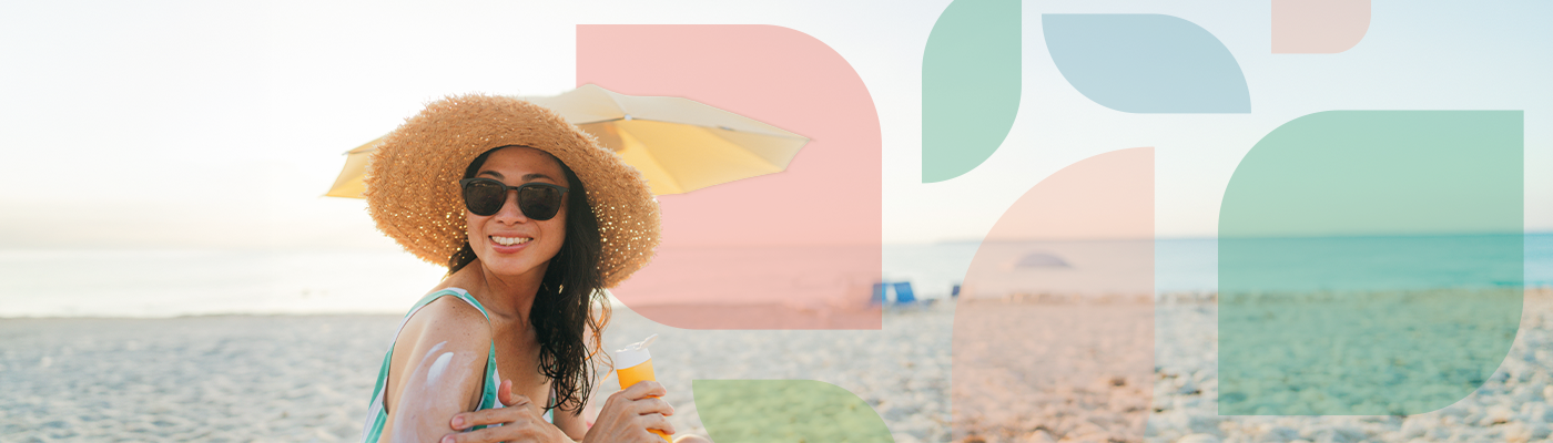 An individual sitting on the beach wearing a sun hat and sunglasses applying sunscreen