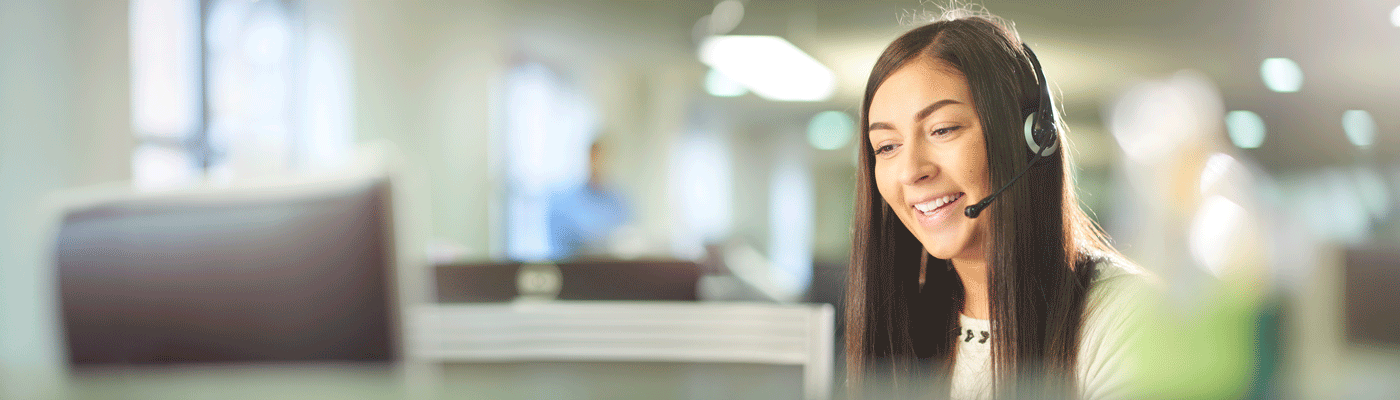 Person wearing a headset seated at a desk in an office environment, with computer workstations and soft overhead lighting in the background